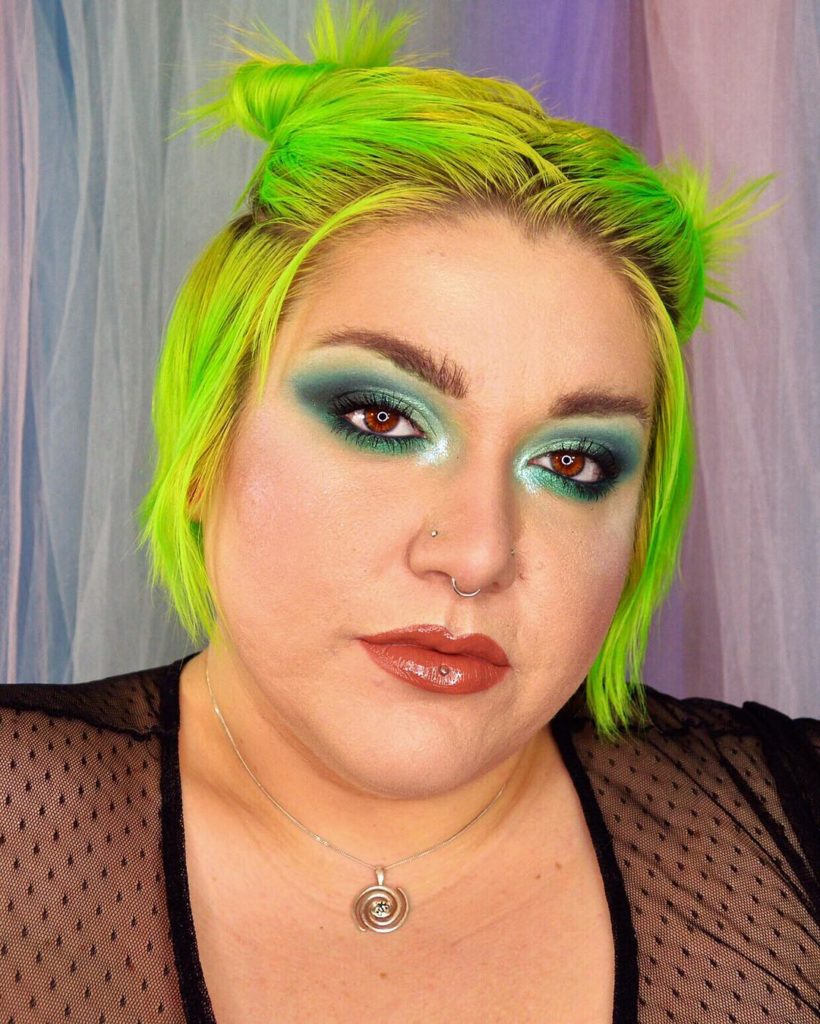 A person with neon green hair with piercings looks at the camera