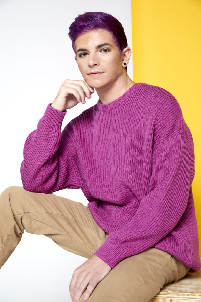 Person with bright purple hair and pink sweater sits in front of yellow background with hand to chin. 