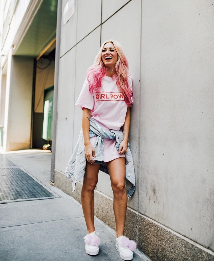 Person with Vibrant and Pastel Pink hair wears a pink shirt and smiles leaning against the wall