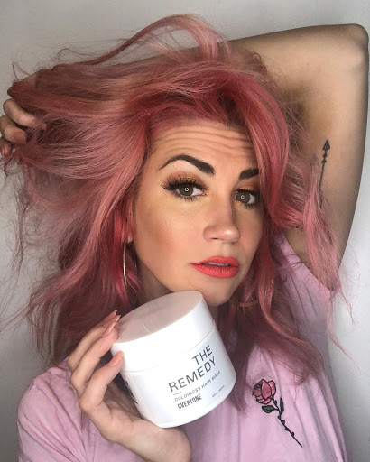 Person with diluted rose gold hair holds a container of The Remedy and looks at the camera