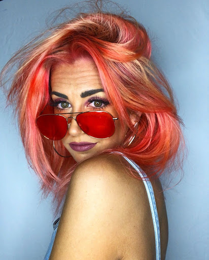 Person with coral and pastel pink hair and wearing sunglasses looks at camera. 