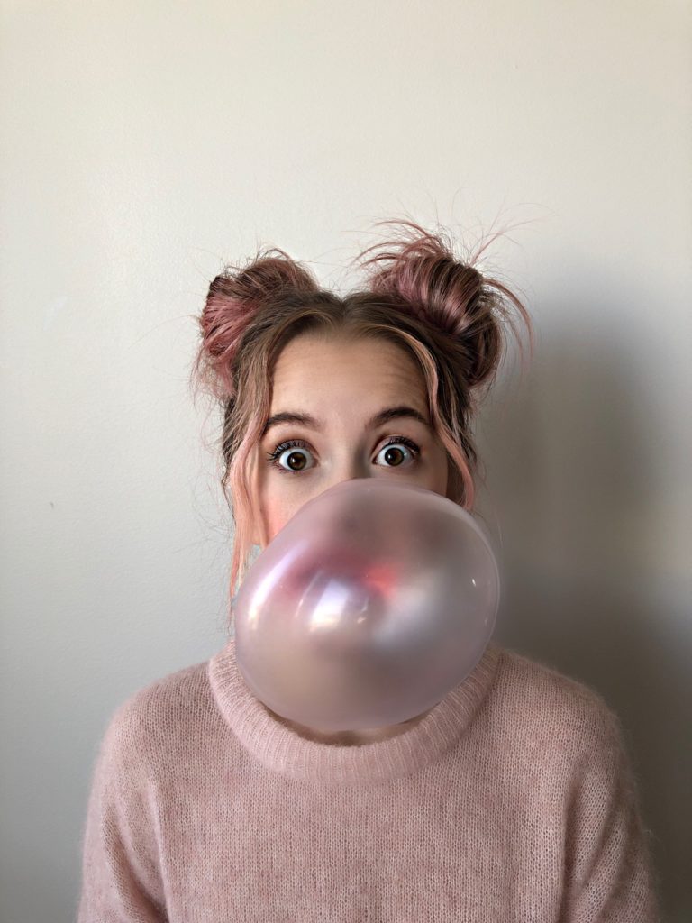 Person with Rose Gold and dark blonde hair with their hair in two top buns. They are blowing a bubble. 
